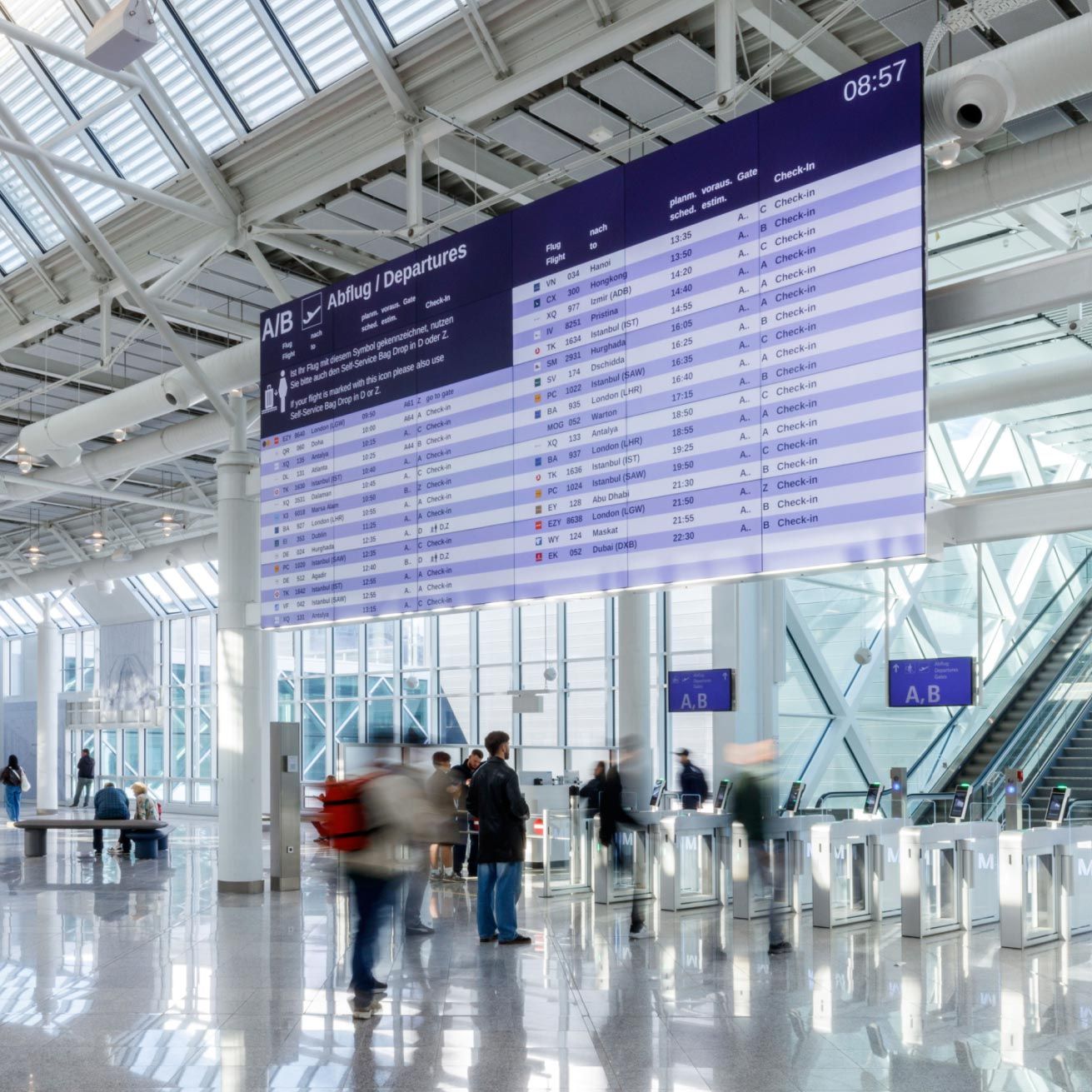 Light-filled departures hall at Munich Airport with a glass roof and glass façade. Beneath a large digital departures board, travellers with luggage pass through a self-service area with check-in and bag-drop machines.