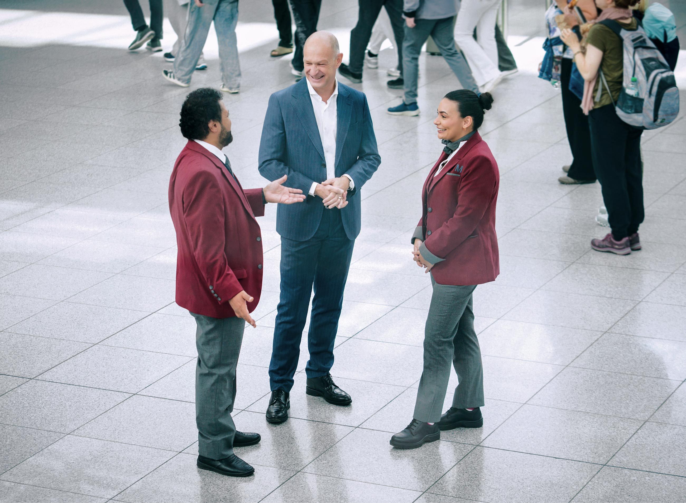 Jost Lammers in conversation with two Munich Airport employees in service uniforms in a busy terminal area; travellers pass by in the background.
