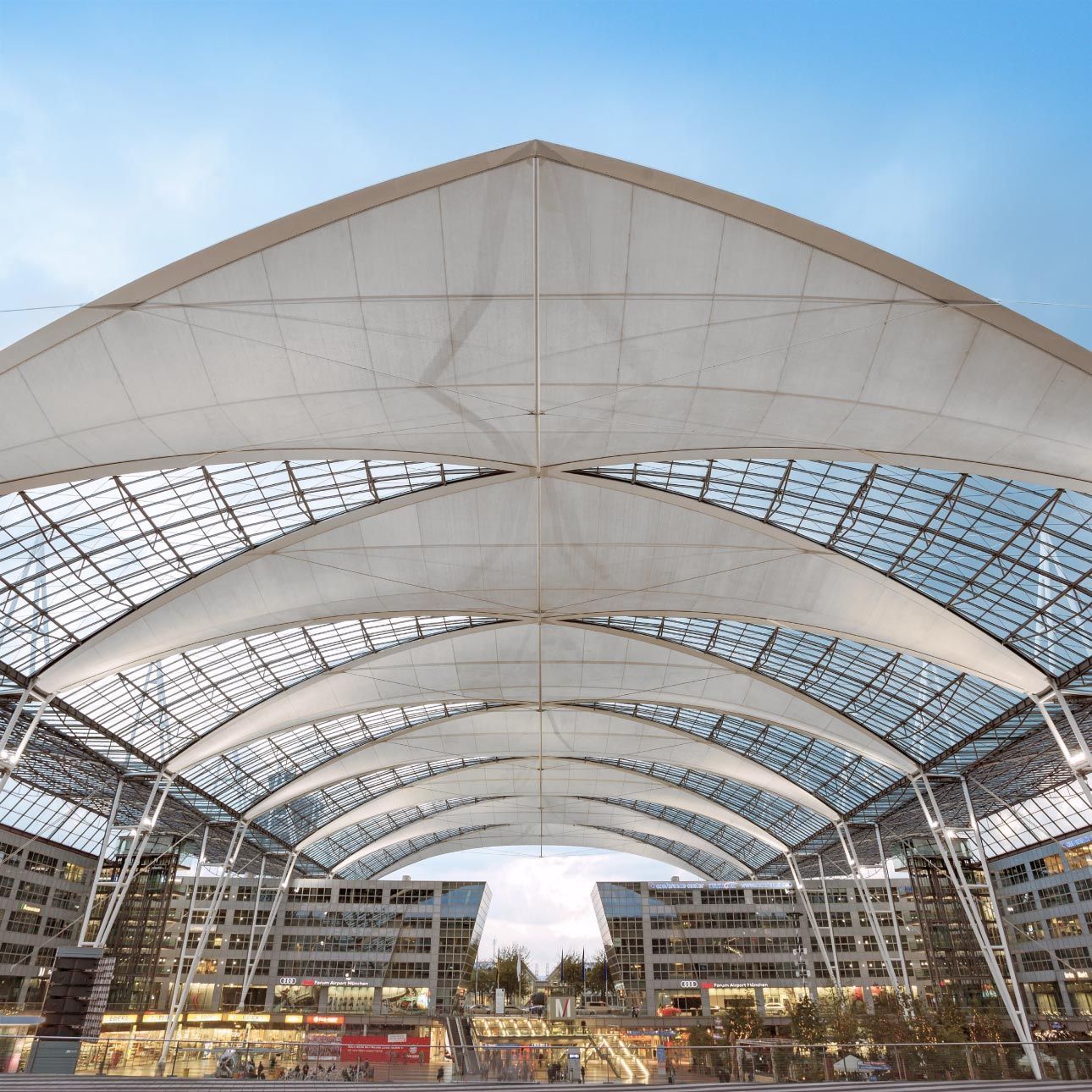 The large, tent-like membrane-and-glass roof over the central plaza of Munich Airport, spanning in wide arches; below it buildings and people, above it a blue sky.