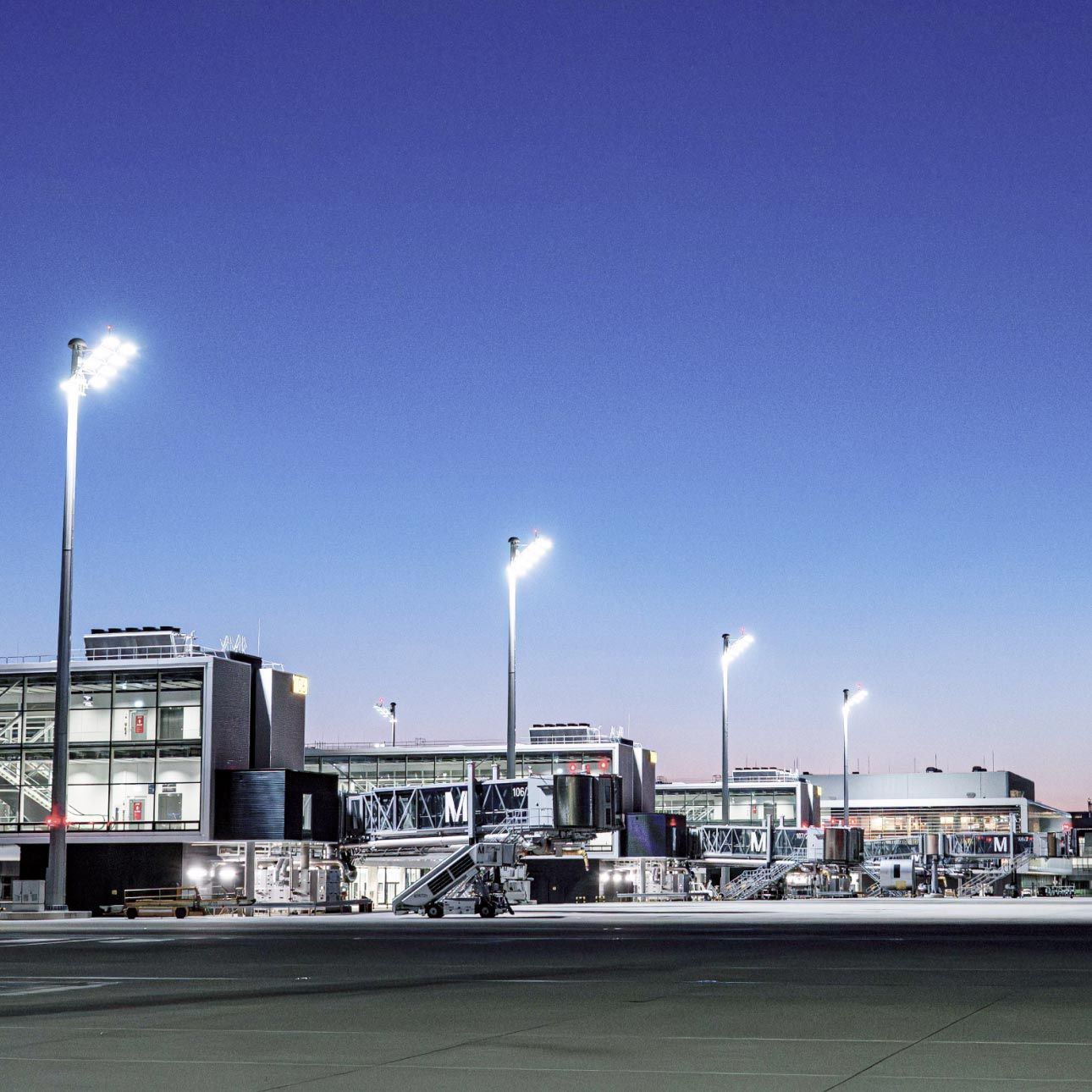 Terminal buildings at Munich Airport with passenger boarding bridges on the apron at dusk, lit by tall floodlight masts against a deep blue evening sky.