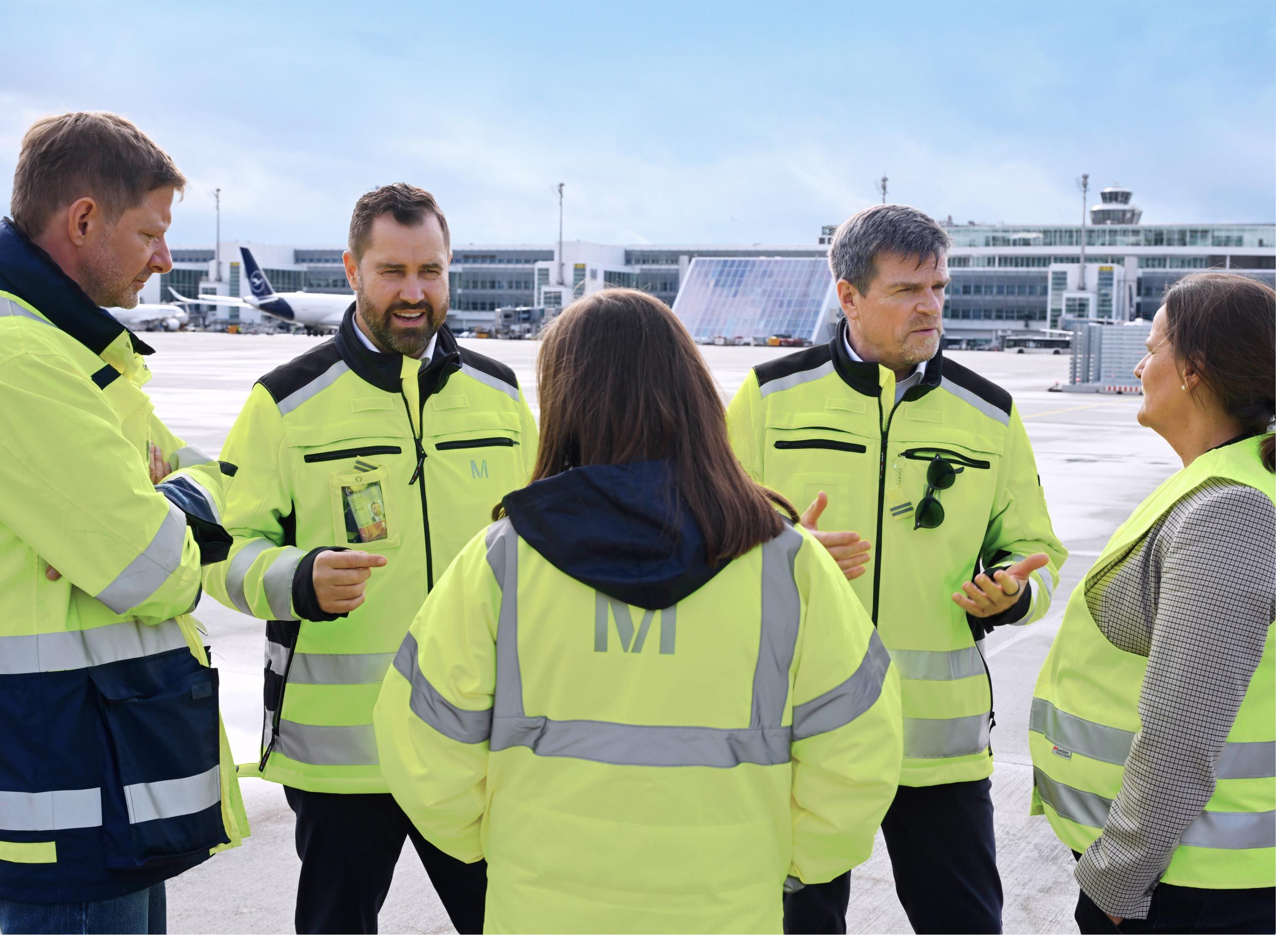 Thomas Hoff Andersson, Managing Director Aviation and Operation, in conversation with four Munich Airport employees in yellow high-visibility jackets with the “M” logo on the apron; in the background terminal buildings, the control tower and an aircraft at the gate.