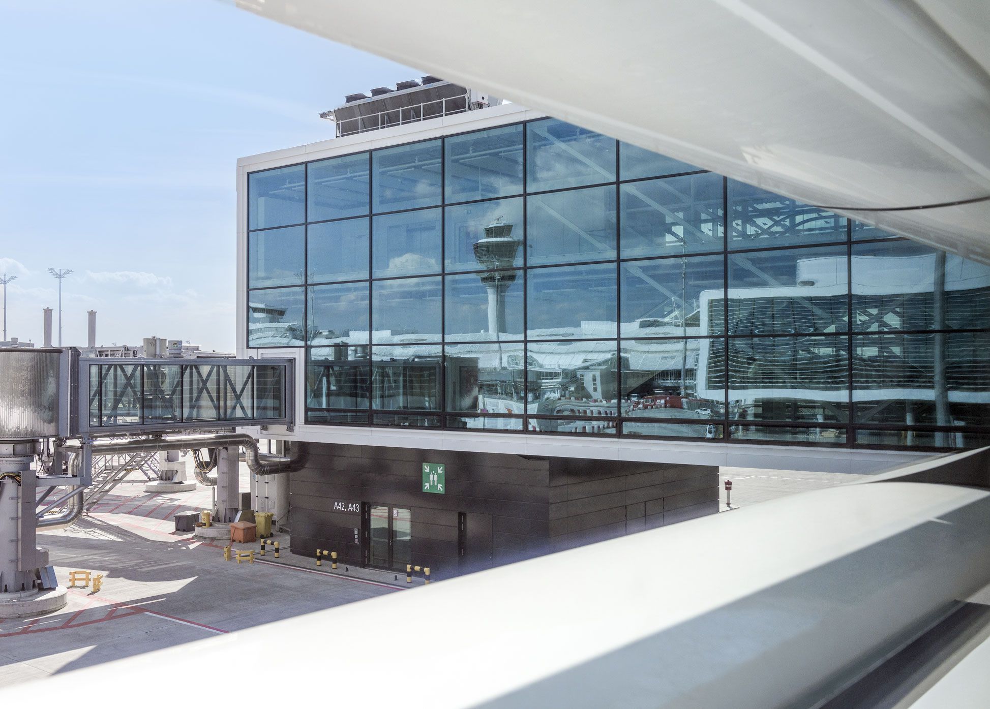 Glass façade of a terminal building at Munich Airport with an attached passenger boarding bridge to gates A42 and A43; the control tower is reflected in the glass front.