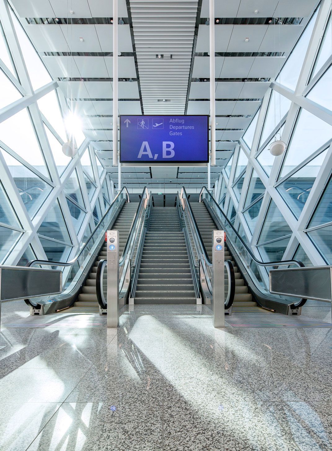 Escalators in a light-filled, glazed terminal hall at Munich Airport, with a sign above reading “Abflug / Departures / Gates A, B”.