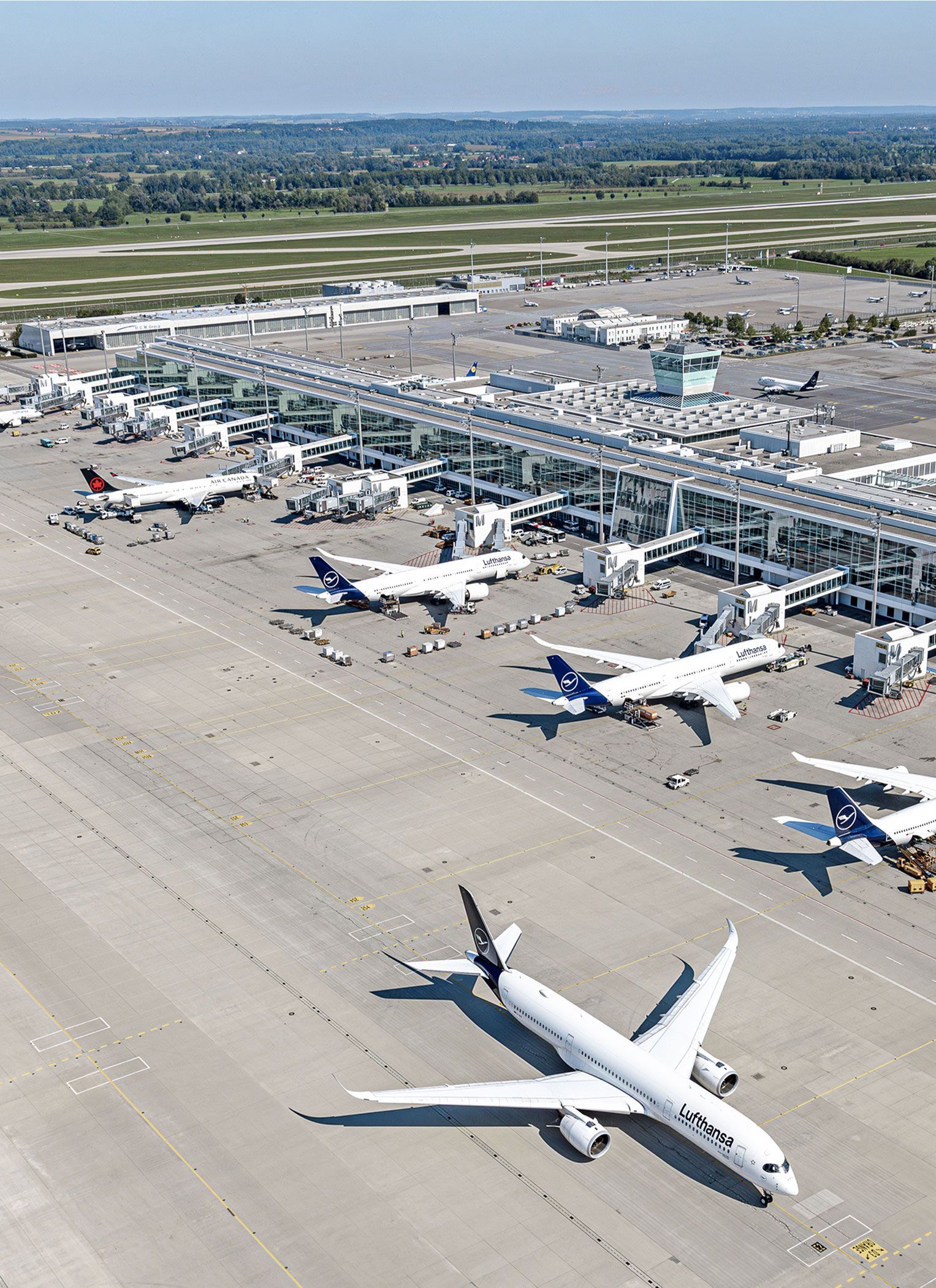 Aerial view of Munich Airport with terminal, apron and control tower; several passenger aircraft stand at the boarding bridges and a Lufthansa aircraft taxis across the apron.