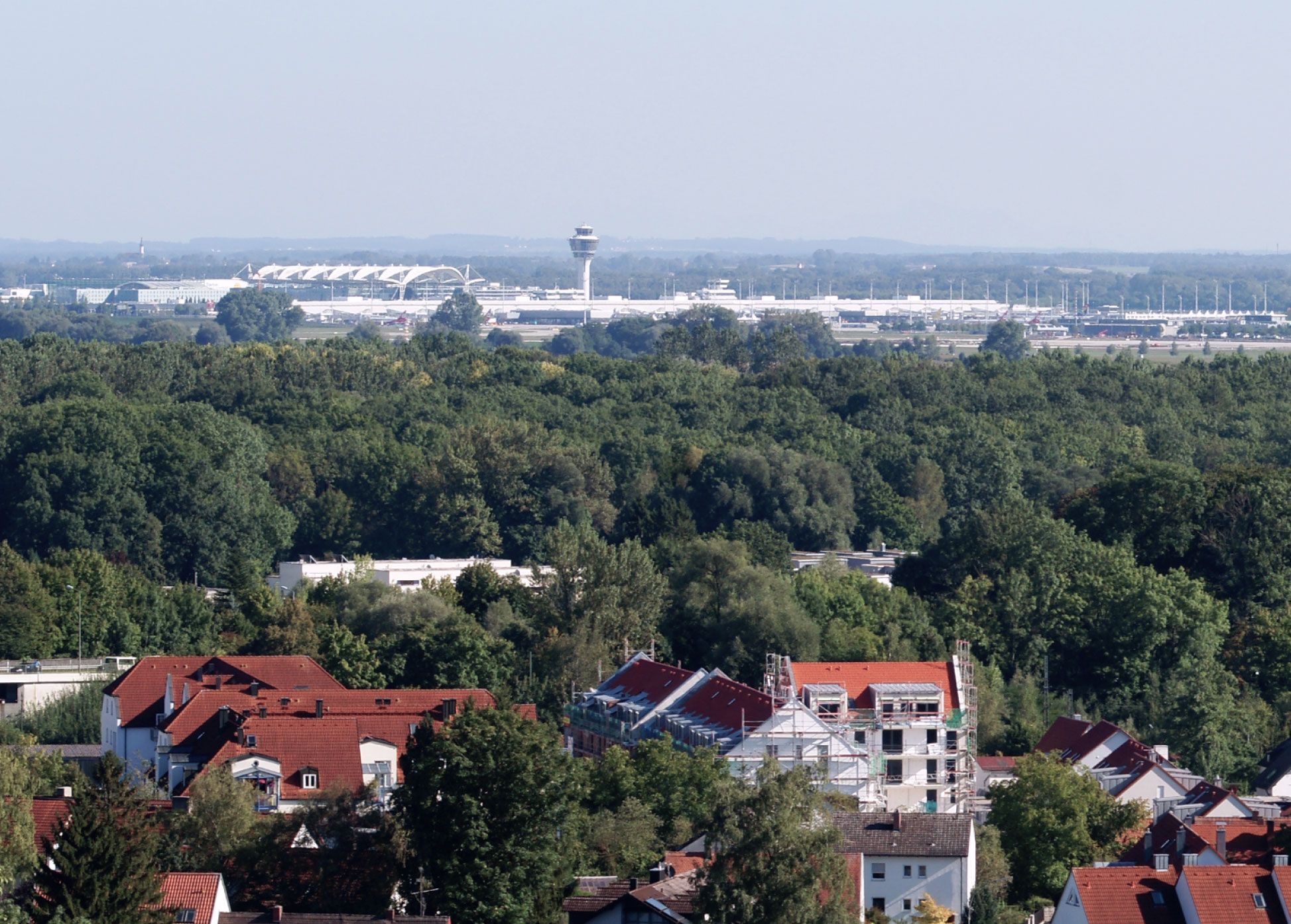 View over a wooded town with residential houses towards Munich Airport on the horizon, with its control tower and terminal roof.